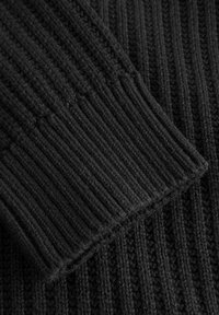 Detail eines schwarzen Strickpullovers mit geripptem Muster und unterschiedlichen Maschenstrukturen, mit einem umgeschlagenen Bündchen in einem dunkleren Schwarzton.