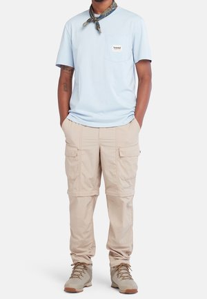 Man wearing light blue short-sleeve shirt, beige cargo pants with zip-off legs, beige hiking shoes, and patterned neck scarf, hands in pockets.