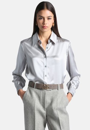 Femme portant une chemise en satin argenté à manches longues, un pantalon gris à chevrons et une ceinture taupe avec une boucle dorée, debout les mains dans les poches.