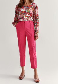 Blouse à manches longues florale en rose, avec des motifs éclatants. Associée à un pantalon tailleur taille haute rose et des sandales à talons roses clairs.