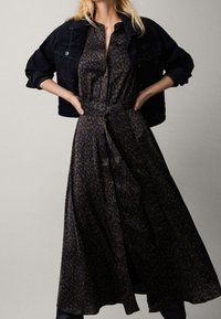 Robe longue en imprimé léopard marron en tissu lisse, avec un lien à la taille, un col, et une silhouette fluide, associée à une veste en denim noire.