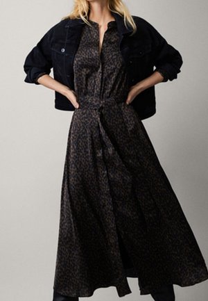 Robe longue en imprimé léopard marron en tissu lisse, avec un lien à la taille, un col, et une silhouette fluide, associée à une veste en denim noire.