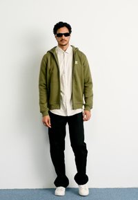 Sweatshirt verde-oliva com fecho, sobre uma camisa bege, combinado com calças pretas e ténis brancos. Modelo a usar óculos de sol pretos, de pé contra uma parede branca.