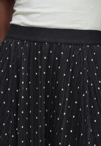 Gros plan d'une jupe plissée noire à petits pois blancs avec une ceinture noire unie portée avec un haut blanc.