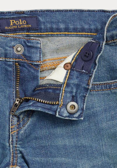 Jeans in denim con zip, caratterizzati da un tessuto blu scuro, elementi in metallo rame e grigio, e cuciture gialle a contrasto sulle cuciture.