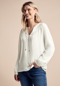 Witte blouse met lange mouwen, een V-hals en trekkoorddetails, met geplooide textuur langs de schouders. Gecombineerd met een blauwe jeans.