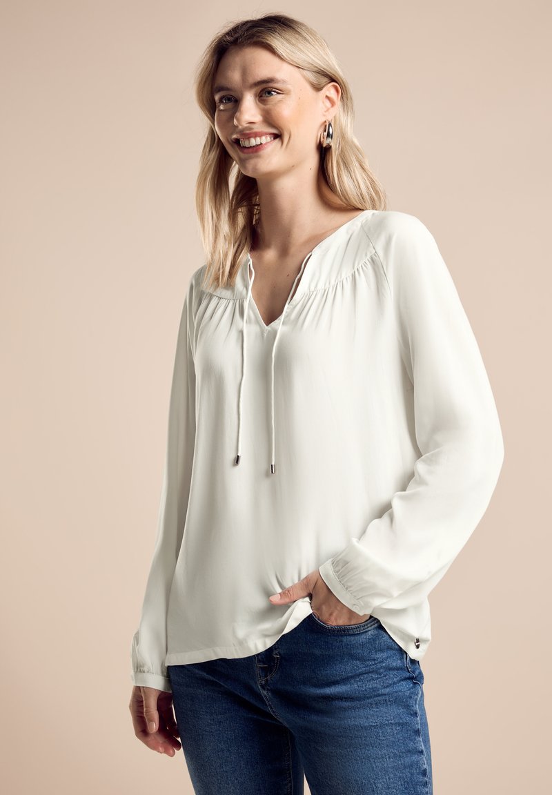 Witte blouse met lange mouwen, een V-hals en trekkoorddetails, met geplooide textuur langs de schouders. Gecombineerd met een blauwe jeans.