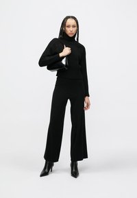 Crni pleteni džemper s visokim izrezom i izraženim uzorkom, upareni sa širokim crnim hlačama i šiljastim crnim gležnjačama. Drži crnu torbicu.