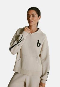 Beige hættetrøje i blødt stof med en forkort lomme, ribbet kant og sorte broderede detaljer på ærmerne og brystet.