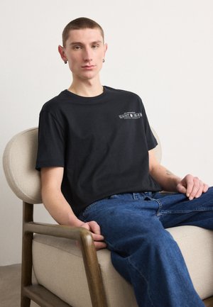 T-shirt in cotone nero con maniche corte, caratterizzato da un logo bianco sul petto. Il modello è seduto su una sedia imbottita in tessuto, indossando jeans di denim blu.
