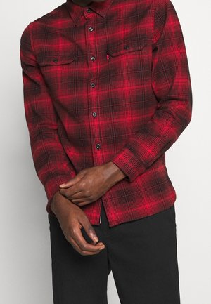 Mand iført rød og sort ternet skjorte med knapper og lange ærmer samt sorte bukser, med armene krydsede foran livet.