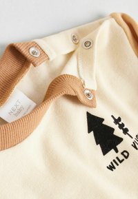 Krèmefarvet babyshirt med brun ribbet krave, trykknaplukninger og sort trægrafik med teksten "WILD VIBES". Blød stofstruktur.