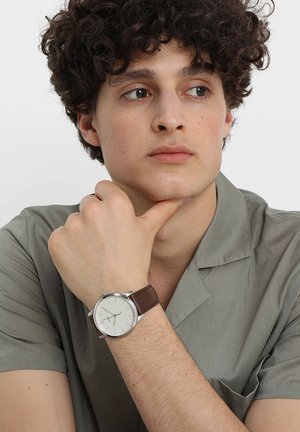 Giovane uomo con capelli ricci che poggia il mento sulla mano, indossa una camicia grigia e un orologio da polso argentato con cinturino in pelle marrone, guarda di lato.