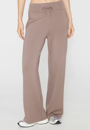 Person trägt lockere taupefarbene Hose mit Kordelzug in der Taille und silberne Sneakers, steht vor einem schlichten hellen Hintergrund.