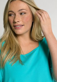 Tyrkysový top s krátkým rukávem vyrobený z měkké tkaniny, s volným střihem a kulatým výstřihem. Vlasy spadají přes ramena, pleť má světlé pihy.