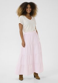 Jupe maxi rayée rose clair et blanche avec un design en étages, taille élastique, associée à un t-shirt crème à col en V à manches courtes et des bottes brunes à la cheville.