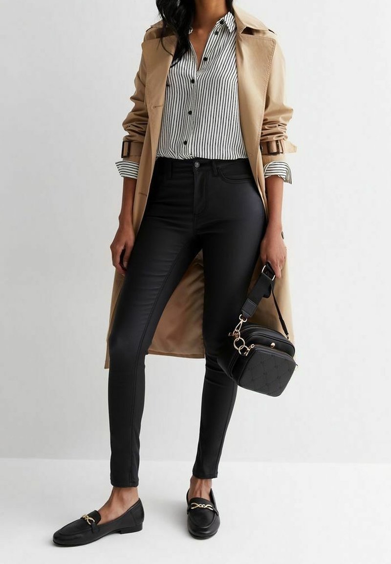 Femme portant un trench-coat beige, pantalon skinny noir, chemise rayée, mocassins noirs, tenant un petit sac à main noir avec des détails en chaîne dorée.