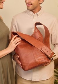 Borsa a spalla in pelle marrone con un design ampio e morbido, tracolla spessa regolabile e dettagli del logo impressi in modo sottile. Texture liscia e colore caldo.