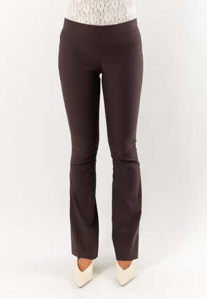 Donna che indossa pantaloni aderenti marrone scuro e scarpe bianche con tacco alto e punta, in piedi su sfondo bianco.