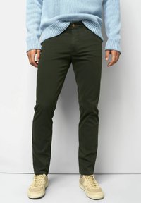 Person in dunkelgrünen Slim-Fit-Hosen, leicht blauer Strickpullover und beigen Sneakern, der vor einem schlichten hellen Hintergrund steht.