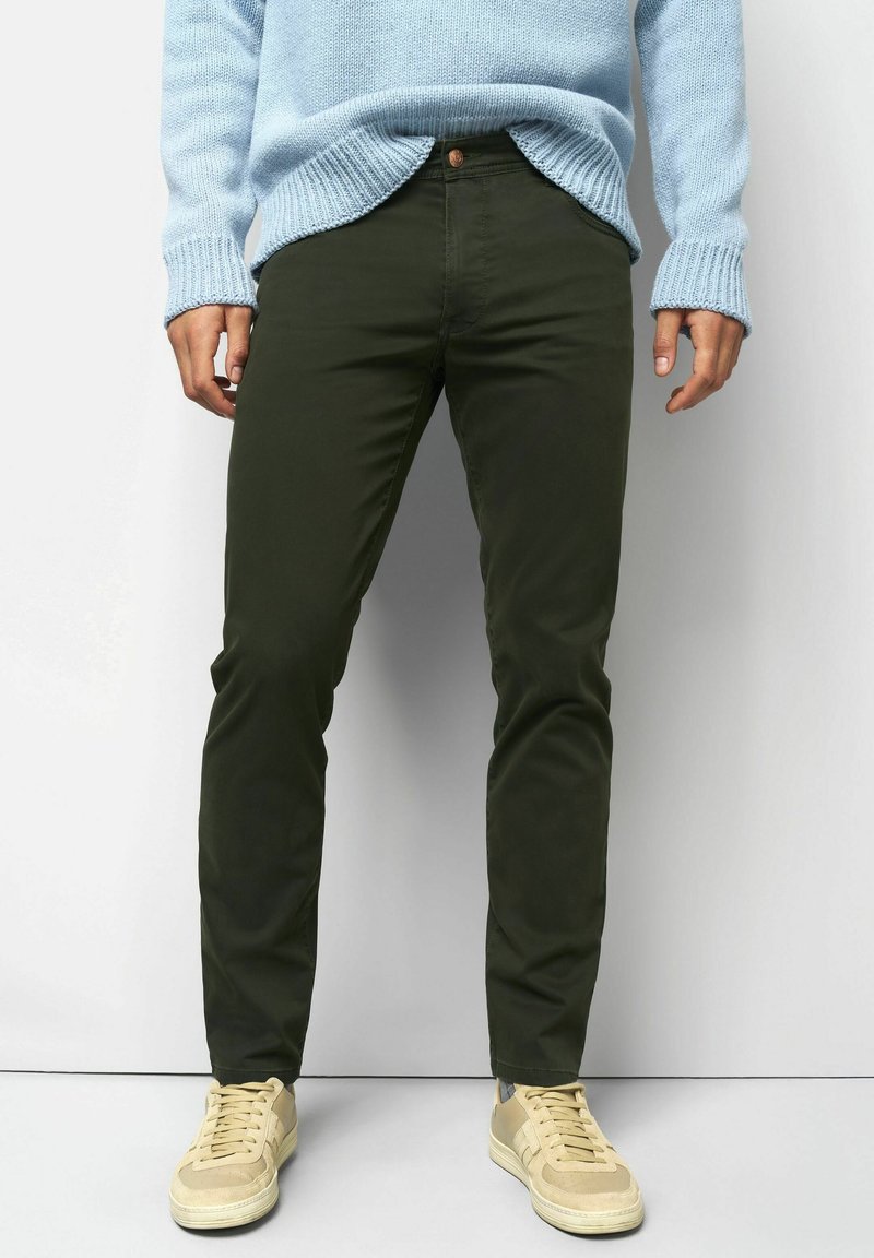 Person in dunkelgrünen Slim-Fit-Hosen, leicht blauer Strickpullover und beigen Sneakern, der vor einem schlichten hellen Hintergrund steht.