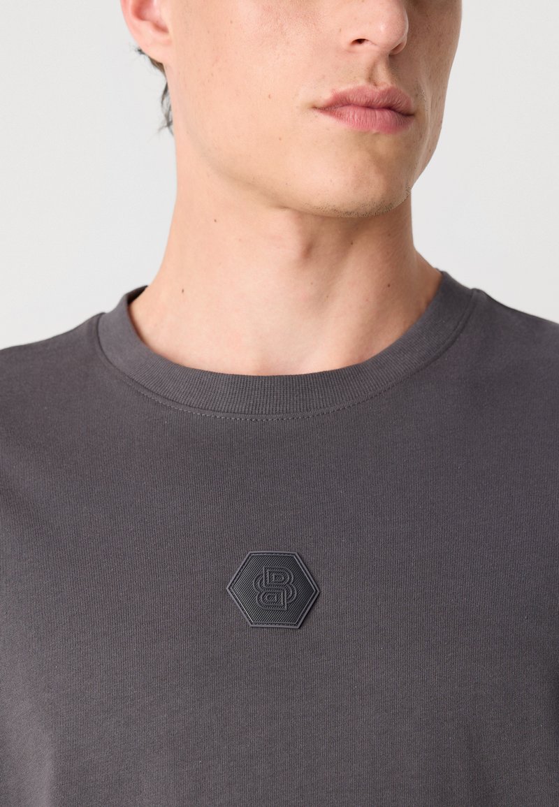 T-shirt gris foncé à col rond, arborant un patch logo hexagonal texturé sur la poitrine. Tissu en coton lisse avec une coupe ajustée.