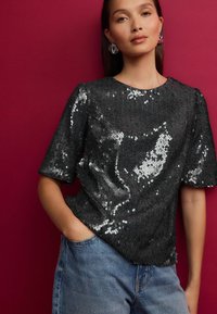 Haut noir pailleté à manches courtes, col rond et coupe légèrement ample, associé à un jean en denim bleu clair. Fond uni.