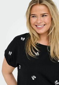 Svart kortärmad topp med vita blommönsterbroderier vid axlarna. Slätt tyg med en avslappnad passform och rund halsringning.