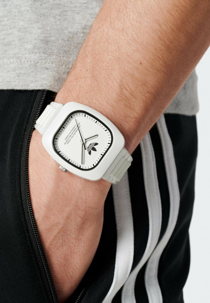 Valge ruudukujuline kell tekstureeritud rihmaga, hõbedase sihverplaadi, mustade märkide ja logoga keskel. Kantud randmel.