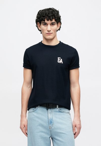 Μπλε ναυτικό κοντομάνικο T-shirt με ριμπ υφή και μικρό λογότυπο "EA" σε ανοιχτό ροζ στο αριστερό μέρος του στήθους, σε συνδυασμό με ανοιχτό τζιν.