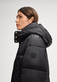 Giubbotto imbottito nero con cappuccio, caratterizzato da imbottitura a cucitura, dettagli in hardware dorato e un patch logo circolare sulla manica.