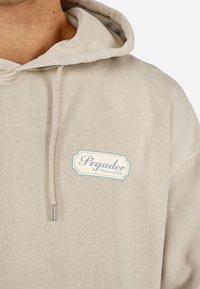 Felpa color crema realizzata in tessuto morbido, con un'etichetta logo con testo blu e rosa sull'area sinistra del petto. Coulisse sul cappuccio.