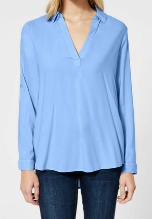 Vrouw draagt een hemelsblauwe blouse met lange mouwen en een open kraag, en donkerblauwe jeans, staand tegen een effen lichte achtergrond.
