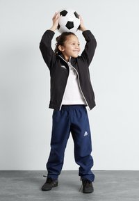 Zwarte zip-up jack, witte t-shirt, marineblauwe sportbroeken met elastische tailleband, met een zwart-wit voetbal boven het hoofd. Zwarte schoenen.