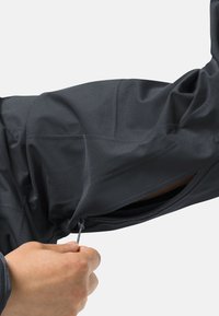 Zwarte stoffen jas met een ritszak op de arm. Het materiaal lijkt lichtgewicht en textuur te hebben, met een aansluitend ontwerp.