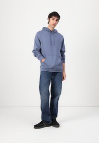 Felpa in cotone blu con tasca frontale e cappuccio con cordoncino, abbinata a jeans blu e sneakers nere. Design semplice con branding minimo.