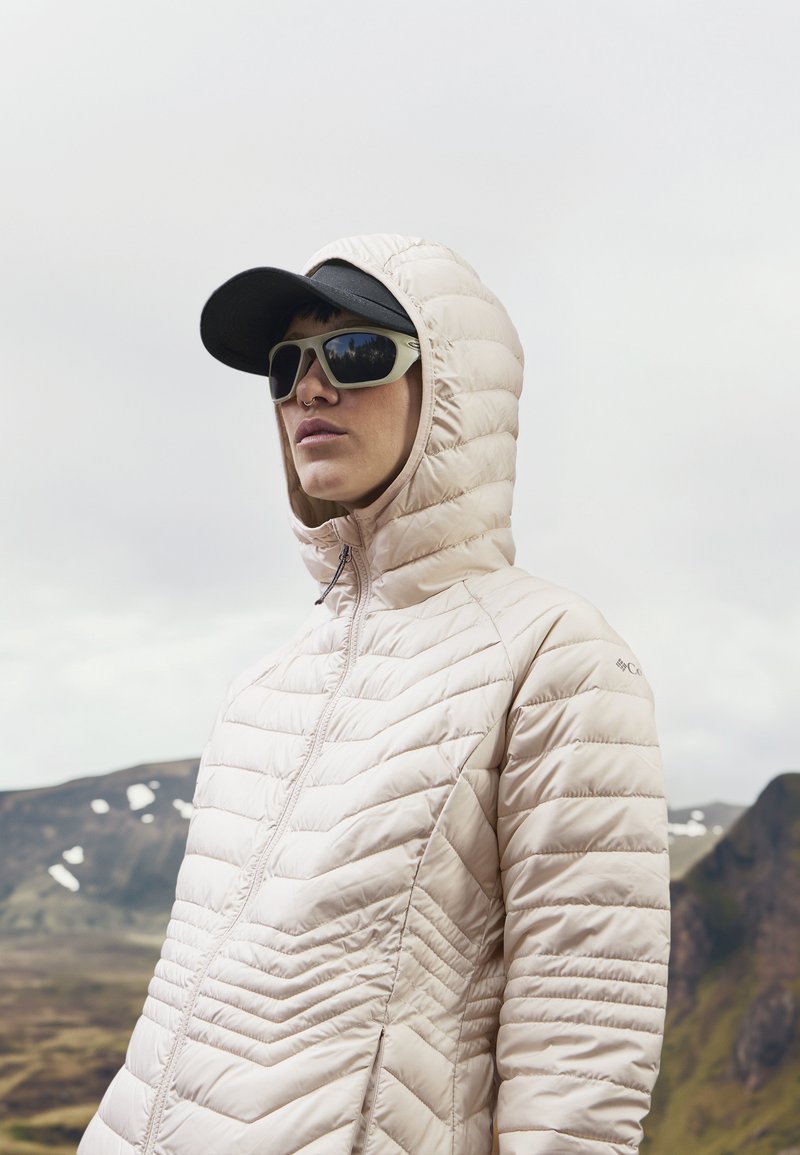 Lichtbeige pufferjack met een geregen patroon, vaste capuchon, volledige ritssluiting, gedragen onder een zwarte pet en zonnebril, tegen een bergachtig landschap.