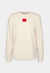 Sudadera beige claro con mangas largas, puños acanalados y una etiqueta de logo cuadrada roja en el pecho. Textura de tela suave y diseño clásico de cuello redondo.