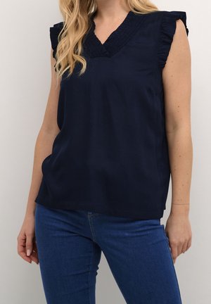 Femme portant un chemisier sans manches bleu marine à volants avec un col en V et un jean bleu sur un fond uni.