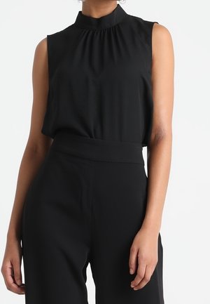 Czarna bluzka bez rękawów z wysokim kołnierzem w połączeniu z czarnymi spodniami z wysokim stanem na stojącej osobie na białym tle.
