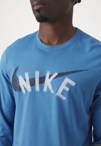 Muž v modré košili s dlouhým rukávem, na které je vybledlý černý logo Nike swoosh a bílé nápis "NIKE" na hrudi, na jednoduchém světlém pozadí.