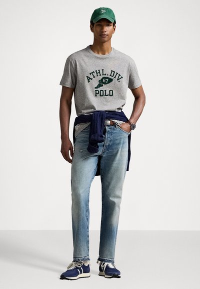 Grijze katoenen t-shirt met de opdruk "ATHL.DIV 67 POLO", blauwe spijkerbroek, marineblauwe trui om de taille geknoopt en blauw/witte sportschoenen.
