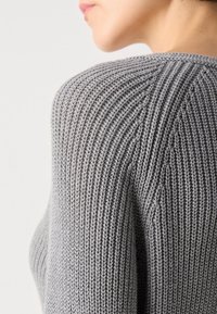 Gros plan sur l'épaule et le haut du bras d'un pull gris en tricot porté par une personne, montrant une texture côtelée détaillée.