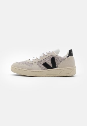 Baskets beige clair avec tissu texturé, logo noir en forme de V sur le côté, lacets blancs et semelle en caoutchouc beige sur fond blanc.