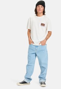 Camiseta blanca de algodón con diseño gráfico en el bolsillo, combinada con pantalones vaqueros de denim azul claro y zapatillas negras con suelas blancas. Lleva puesta una gorra negra.