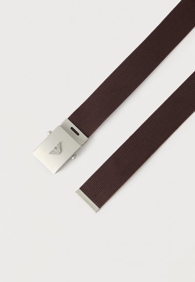 Ζώνη από σκουρόχρωμο καφέ υφαντό ύφασμα με ασημένια ορθογώνια αγκράφα που φέρει ανάγλυφο έμβλημα αετού και ασημένιο μεταλλικό άκρο στο αντίθετο άκρο.