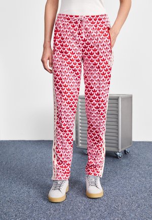 Osoba nosząca czerwono-różowe spodnie dresowe Adidas z wzorem logo trefoil oraz szare trampki, stojąca na niebieskim dywanie.