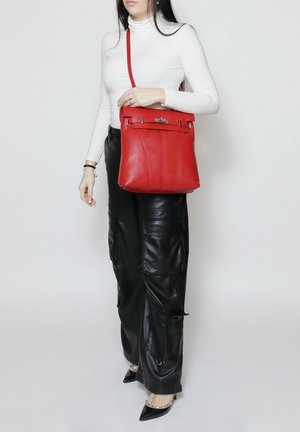 Donna con maglia bianca a maniche lunghe e pantaloni in pelle neri tiene una borsa a tracolla rossa in pelle e indossa décolleté nere con punte a punta e borchie.