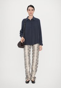 Cămașă lungă cu mânecă navy, croială lejeră, asortată cu pantaloni bej cu imprimeu de șarpe; ținând o geantă de mână din material țesut închis, pantofi negri cu talpă plată, fundal neutru.