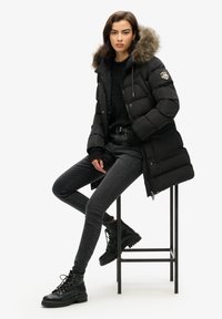 Chaqueta de plumas negra con capucha forrada de piel, con cremallera y cierres de botón, luciendo en un modelo combinado con jeans ajustados negros y botas de cordones.